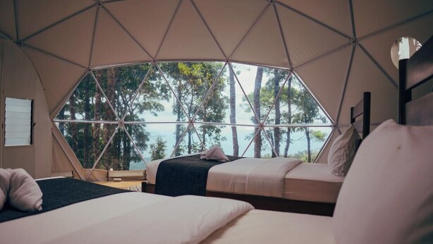 A tent-style room with two beds, a view of trees and water outside, and a window with a geometric pattern. - Matahom Forest Glamping Tent 1 (Mambajao)