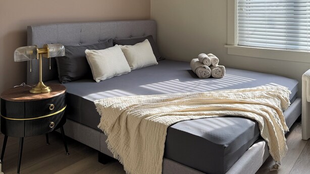 Une chambre moderne avec un lit gris, une table de chevet ronde, un luminaire fixé au mur et une fenêtre avec des stores.