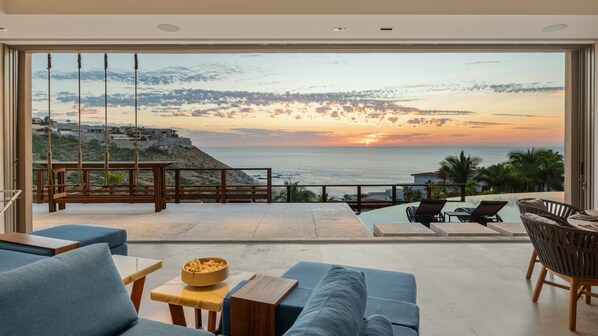 A modern living room with a large glass door leading to a balcony overlooking a coastal sunset.