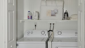 Une buanderie équipée de deux machines à laver à chargement frontal et d’un sèche-linge, d’une étagère pour le linge et le matériel de lessive, et d’une prise de courant.