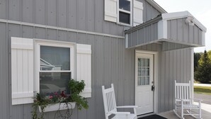 Un bâtiment à l’extérieur gris, avec des volets blancs, une porte d’entrée et deux fauteuils à bascule.