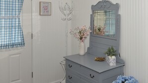 Une pièce avec une commode grise, un vase blanc orné de fleurs, un rideau à carreaux bleus et blancs, et un panneau encadré accroché au mur.