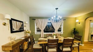 Un comedor con una mesa de madera dispuesta para cuatro personas, rodeada de sillas. Se observa una araña sobre la mesa y una ventana con cortinas.