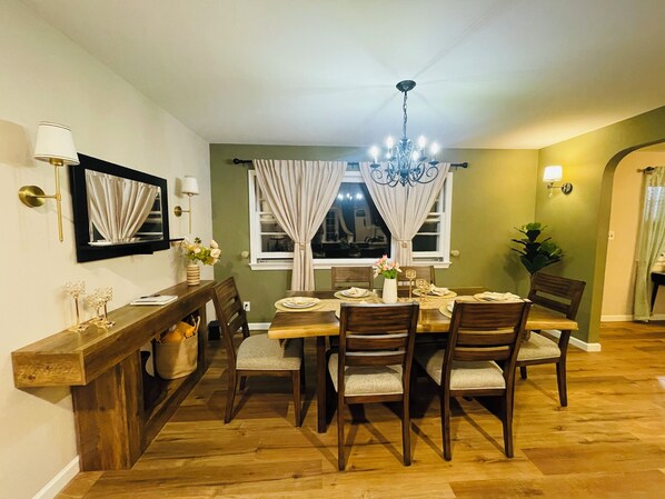 A dining room with a wooden table set for four, surrounded by chairs. There is a chandelier above the table, and a window with curtains.