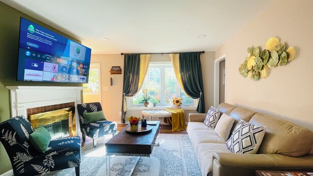 Una sala de estar con chimenea, televisor montado en la pared, sofá, mesa de centro y ventana con cortinas.