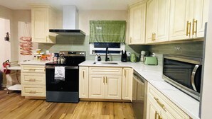 Una cocina con armarios de color claro, un lavavajillas negro, un microondas y una ventana con cortina verde.