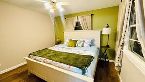 Un dormitorio con cama, ventilador de techo, ventana con cortinas, lámpara de noche y lámpara de pie.