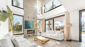 A modern living room with a fireplace, white sofas, a coffee table, and large windows offering a view of the outdoors. - Villa El Paraiso | Cinema Gym Pool Parking (Benahavís)