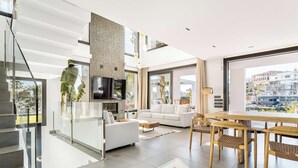 A modern living room with a sofa, coffee table, and dining table. There is a staircase with glass railings and a TV mounted on the wall. - Villa El Paraiso | Cinema Gym Pool Parking (Benahavís)