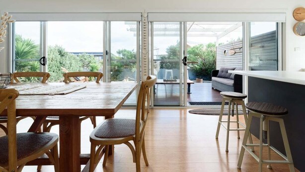 A spacious dining area with a wooden table and chairs, a kitchen area with a bar, and a sliding glass door leading to a patio with outdoor furniture. - Low Head Coastal Haven: Your Seaside Escape (George Town)