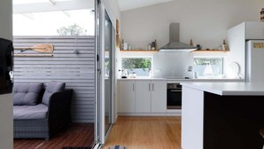 A modern kitchen with white cabinets, a stainless steel range hood, and a built-in oven. There is a sliding glass door leading to a patio area with a wicker sofa and a wooden deck. - Low Head Coastal Haven: Your Seaside Escape (George Town)