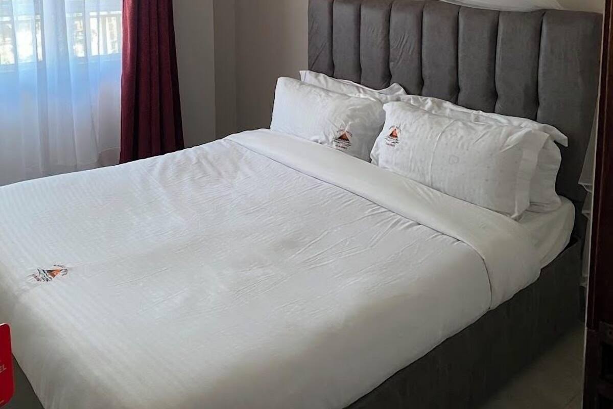 A bed with white linens, a grey upholstered headboard, and a sheer canopy.