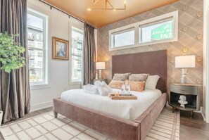 A modern hotel room with a large bed, a nightstand, a lamp, and a view of the city through the window. - Fall in Nashville: 4BR Germantown house, sleeps 12, near Broadway (Nashville)