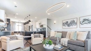 Een moderne woonkamer met een bank, fauteuils en een salontafel. In de achtergrond is een keuken te zien, met een trap naar een ander niveau.