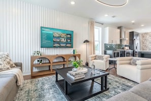 A modern living room with a flat-screen TV, a wooden console table, and a coffee table. There is a kitchen area visible in the background.