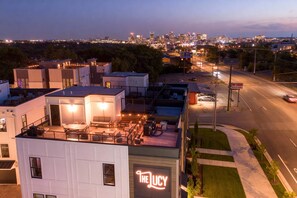 A modern building with a rooftop patio area, a neon sign reading "The Lucy," and a view of a city skyline in the background.
