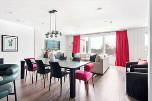 A modern living room with a dining area, featuring a large window with red curtains, a sofa set, and a dining table with chairs.