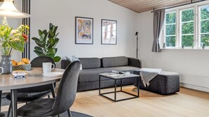 Una sala moderna con un sofá gris, una mesita de centro y un área de comedor con sillas y una mesa. Hay dos cuadros en la pared, una planta en maceta y una ventana con cortinas.