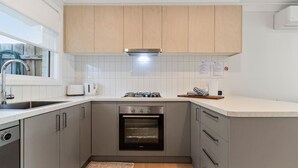 Una cocina compacta con gabinetes grises, horno empotrado, lavamanos de acero inoxidable y ventana con vistas al exterior.