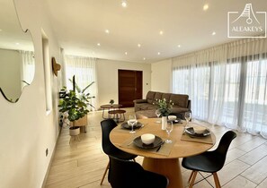 A modern living room with a dining area, featuring a wooden dining table set for four, black chairs, a brown sofa, and a large window with sheer curtains.