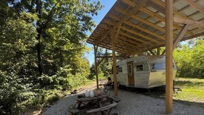 Un càmping amb una taula de picnic coberta, una xemeneia i una caravana estacionada sota una pèrgola de fusta.
