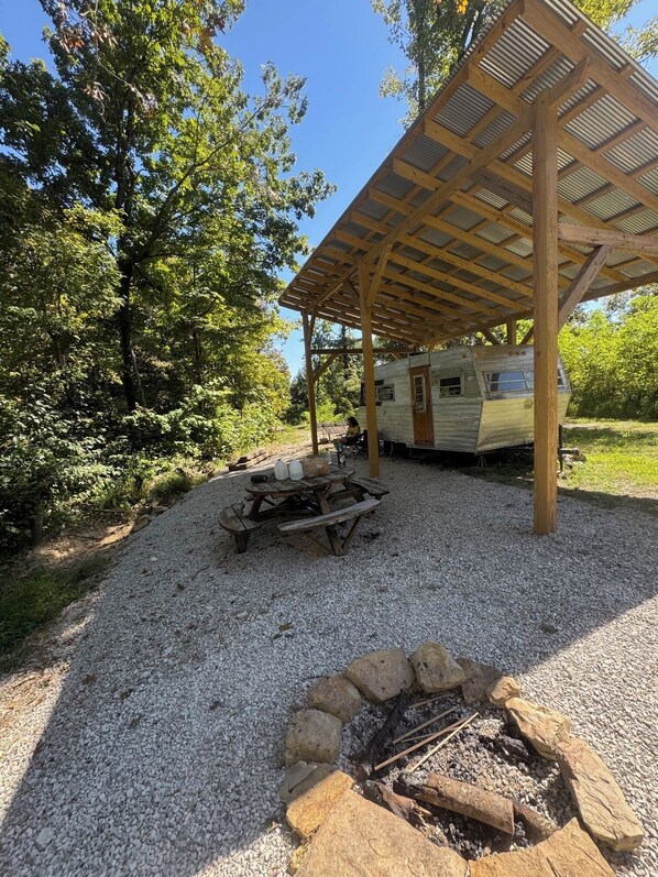 A campsite with a covered picnic table, a fire pit, and a camper parked under a wooden pergola.