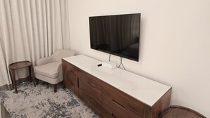 Een moderne hotelkamer met een groot flatscreen aan de muur, een houten dressoir met lades, een beige fauteuil, een klein rond bijzettafeltje en een kamerbreed tapijt op de vloer.