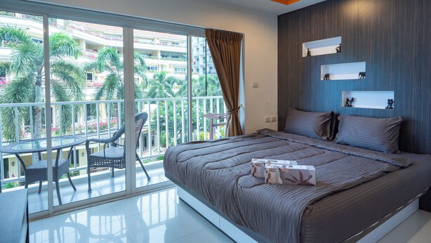 Une chambre avec un grand lit, un balcon avec des chaises et une vue sur les palmiers et les immeubles.