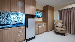 Une cuisine moderne avec des meubles en bois, un four à micro-ondes encastré et un réfrigérateur. Un téléviseur est fixé au mur et un fauteuil beige se trouve dans un coin.