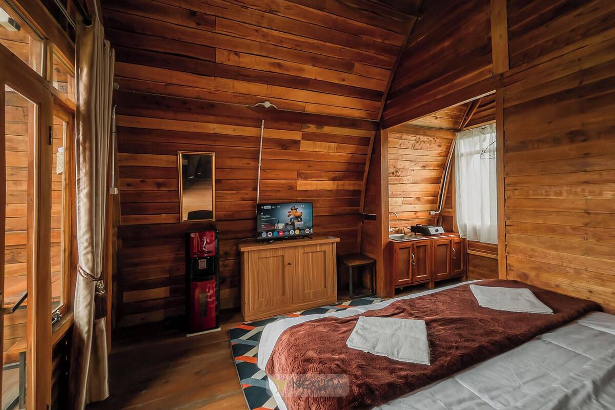Una habitación acogedora tipo cabaña con paredes de madera, una cama con una manta estampada, un televisor sobre un mueble de madera y una pequeña cocina.