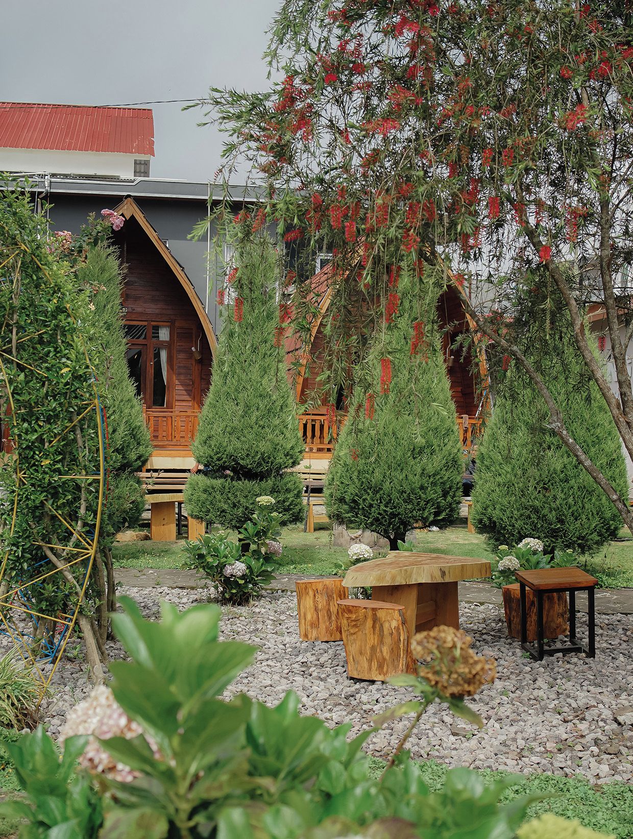 Una cabaña de madera con un porche, rodeada por un jardín con arbustos variados y una pequeña mesa de madera.