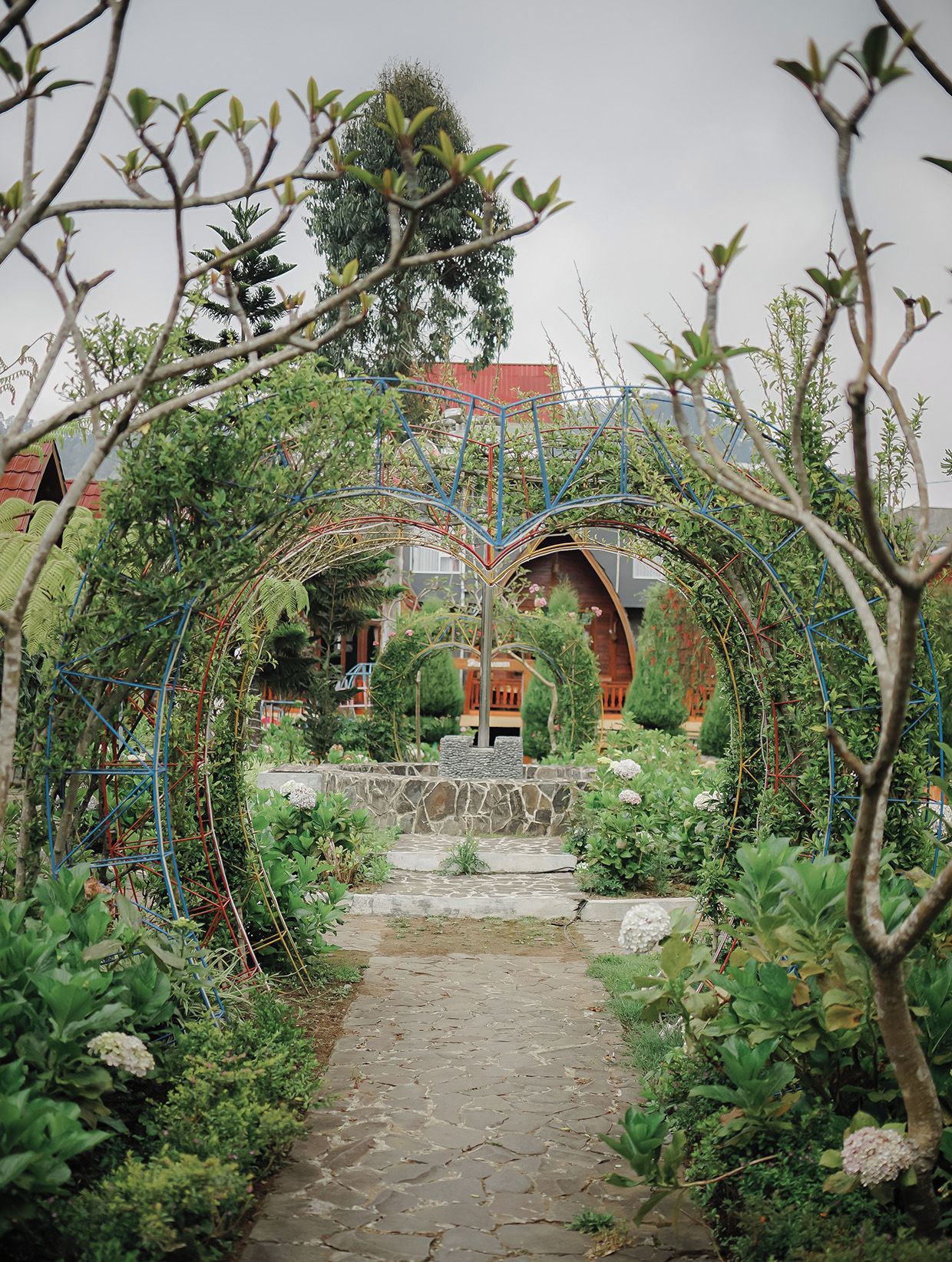 A garden pathway leading to a building with a red roof, surrounded by lush greenery and a decorative metal archway.