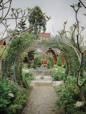 A garden pathway leading to a building with a red roof, surrounded by lush greenery and a decorative metal archway. - Omah Lumbung Nexura Escapes (Dieng Kulon)