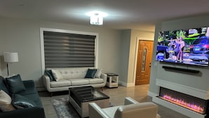 Ein modernes Wohnzimmer mit einer Couch, Sesseln, einem Couchtisch, einem Kamin und einem Fernseher, der eine Nachrichtensendung zeigt.