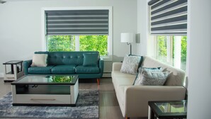 Ein Wohnzimmer mit einem grünen Sofa, einem weißen Sofa, einem Glastisch und einer Lampe. Zwei Fenster mit Jalousien und ein kleiner Tisch mit einer Lampe sind ebenfalls vorhanden.