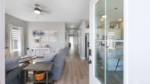 Una cocina moderna con separador de vidrio, una zona de comedor con sofá y mesa, y un pasillo con obras de arte.