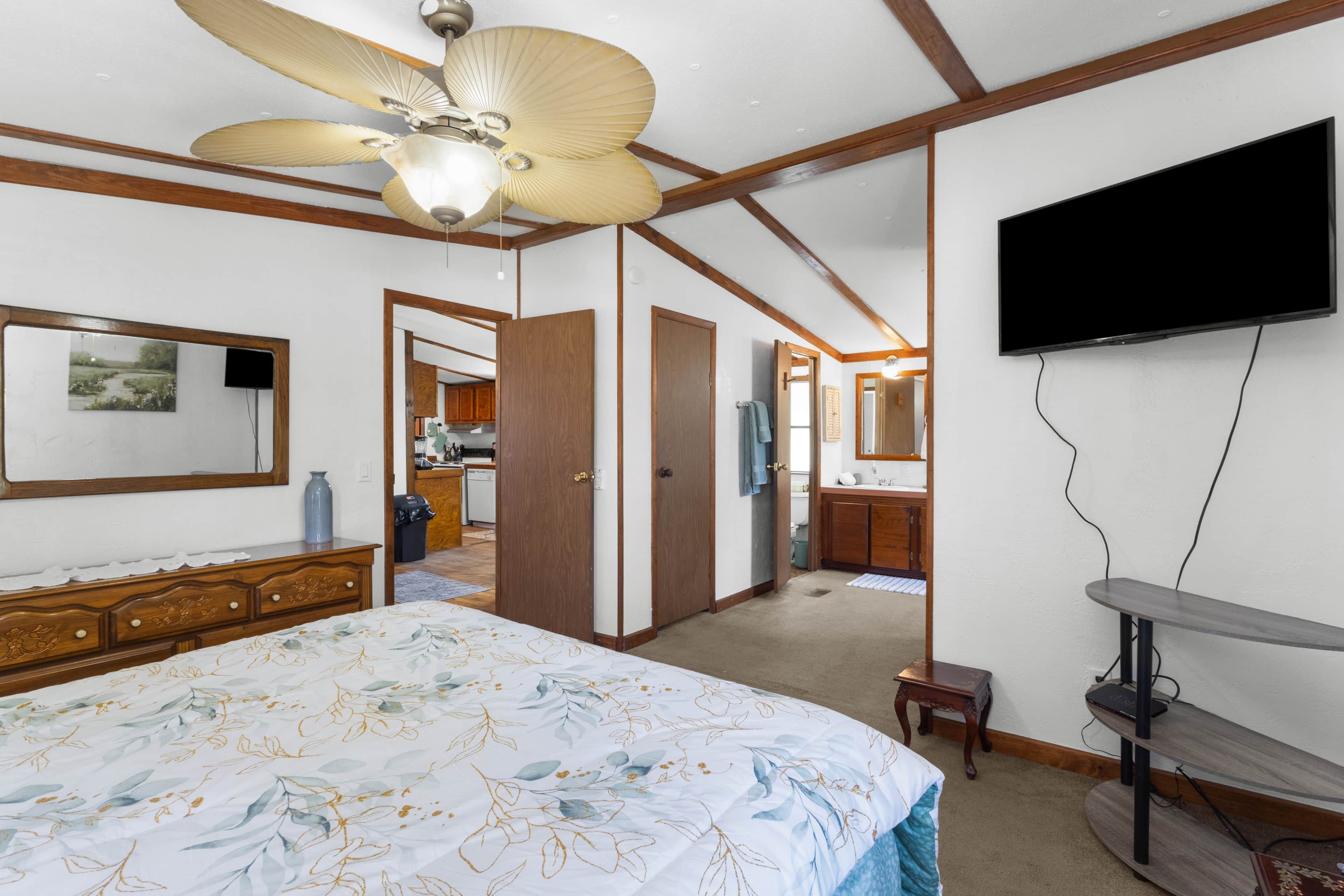 A bedroom with a bed, a ceiling fan, a television, and a view into the kitchen.
