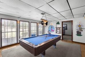A pool table in a room with large windows, a ceiling with a grid pattern, and wall decorations including a compass and a ship.