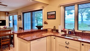 Cocina con encimeras de granito, lavavajillas y fregadero. Hay un comedor con mesa y sillas, y una ventana con vistas a árboles y una masa de agua.