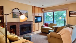 Una sala de estar acogedora con chimenea, un televisor instalado en muebles de madera y un ventanal con vistas a árboles.