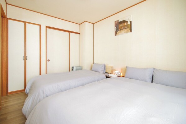 A bedroom with two beds, wooden flooring, a radiator, a bedside table with a lamp, and a framed picture on the wall. - YADOKITO2 / Otaru Hokkaidō (Otaru)