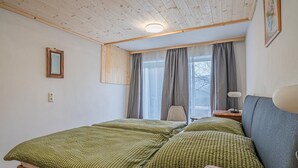 A bedroom with a wooden ceiling, a bed with green bedding, a chair, a desk with a lamp, and a window with curtains. - Quaint holiday home with garden and views (Oberndorf in Tirol)
