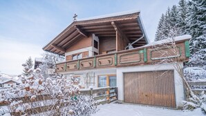 A two-story chalet with a wooden facade, a balcony, and a garage. - Quaint holiday home with garden and views (Oberndorf in Tirol)