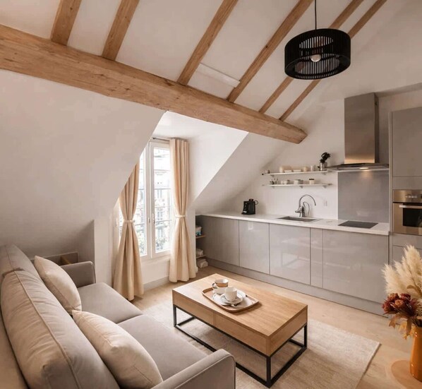 A modern loft apartment with a sloped ceiling, a cozy living area with a sofa and coffee table, a kitchen with a built-in island, and a large window with curtains. - Place des Vosges - Deluxe apartment for 4 (Paris)
