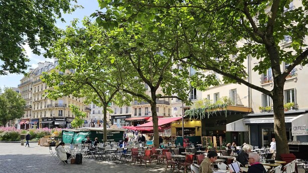 Un café al aire libre con mesas y sillas, rodeado de árboles y edificios.