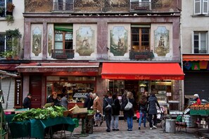 A street with a market stall selling vegetables and a red awning with a sign that reads 'Gastronomia Italiana.'