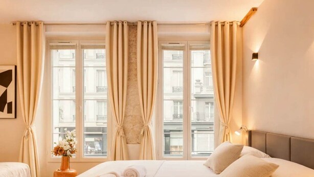 A hotel room with a large bed, two bedside tables, a vase with flowers, and a view of buildings through the windows. - Marais stylish & central studio (Paris)