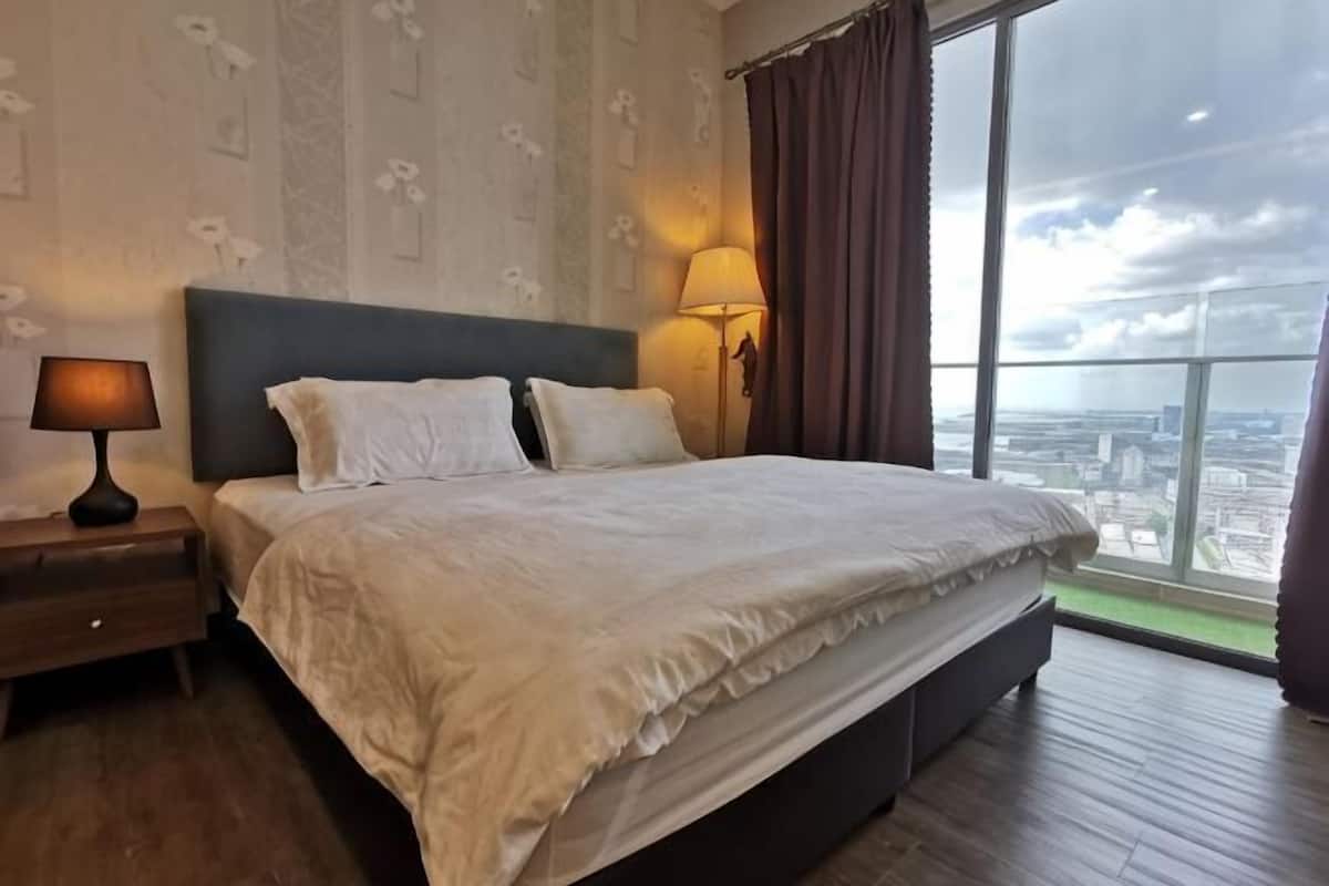 A large bed with a headboard, a bedside table with a lamp, and a view of a cityscape through large windows.