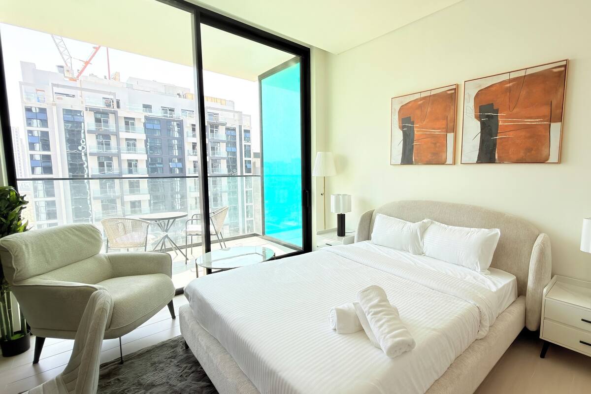 A hotel room with a bed, a chair, a nightstand, and a view of the cityscape through large windows.