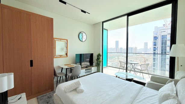 A modern hotel room with a large bed, a desk with a TV, a small balcony with a table and chairs, and a city view. - Premium Studio In Jvc Bazr (Dubai)